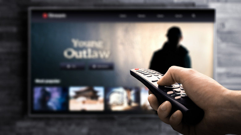 TV out of focus in the background, with foregrounded hand holding a remote pointed at it.