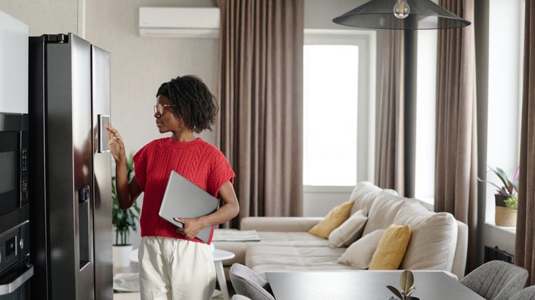 A woman in a red shirt and glasses touches the screen of her smart fridge while holding a laptop.