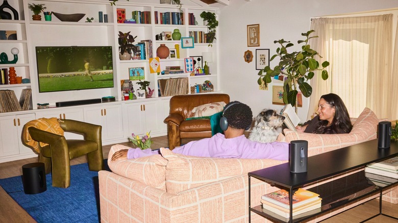 A couple sitting on a couch with a dog in between them, with some Sonos products around the room.