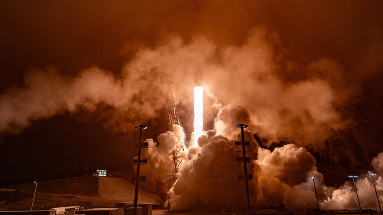 A SpaceX Falcon 9 rocket launches in a red cloud of smoke.