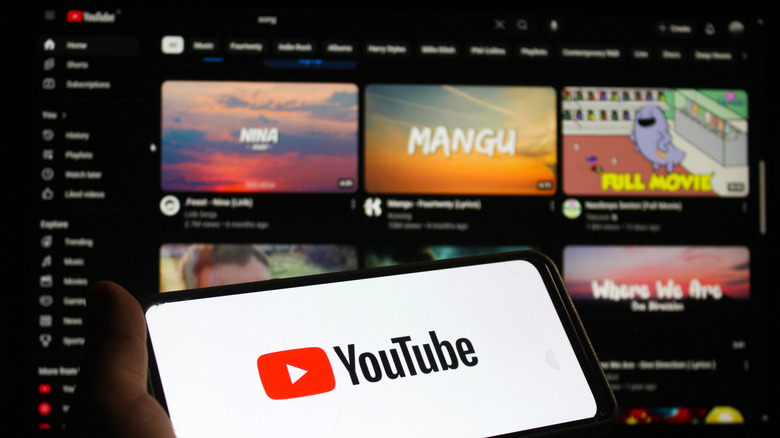 The YouTube logo showing on a smartphone, in front of the homepage on a monitor.