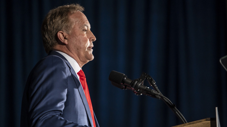 Profile photo of Texas Attorney General Ken Paxton. He's wearing a blue suit and red tie, speaking into a microphone in front of a dark blue curtain.
