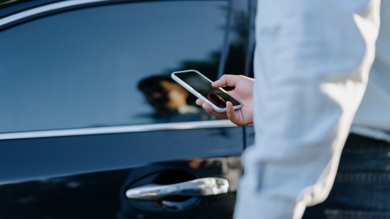 A man in a suit holding a phone in front of his car.