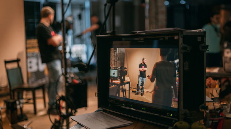 Film production set with a monitor showing a person being recorded on camera while crew members work in the background.