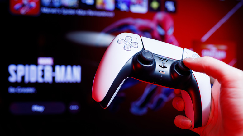 Hand holding up a DualSense controller in front of a TV showing the Spider-Man splash screen.