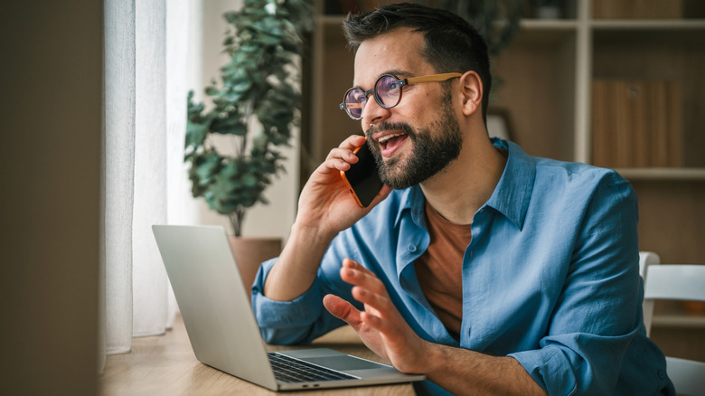 A man wearing glasses, smiling while talking on the phone in front an open laptop.