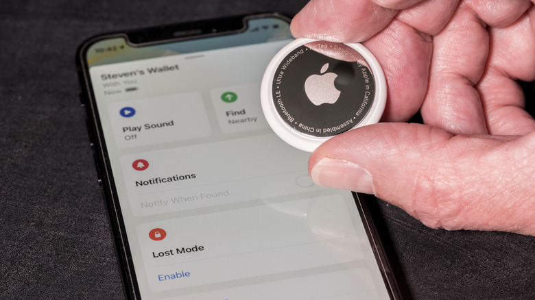 Fingers holding an Apple AirTag in front of an iPhone showing the Find My app.