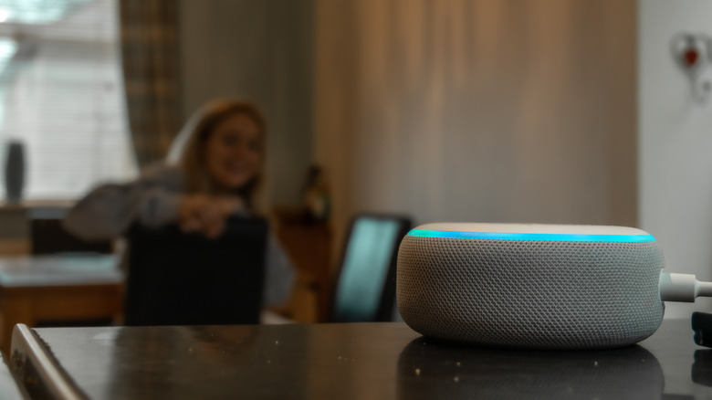 An Amazon Echo Dot sitting on a table in the foreground. In the background a woman is sitting down out of focus.