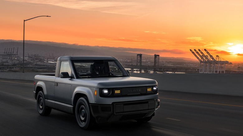 A grey slate truck drives across a bridge at sunset, where a busy port is just visible against the yellowing sky.