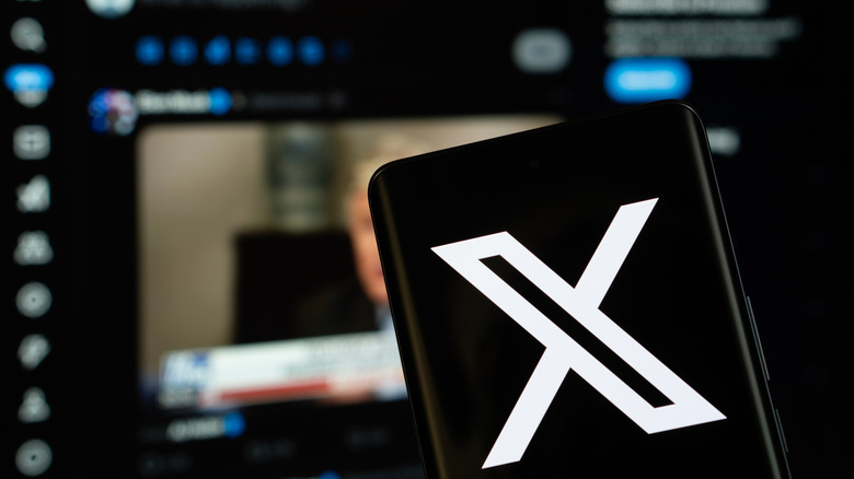 An X logo on a phone with the X desktop interface blurred in the background.