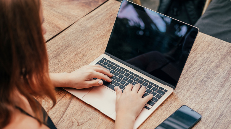 Person using a MacBook on a wooden outdoor table, with reflections visible across the dark screen and a smartphone beside the laptop.