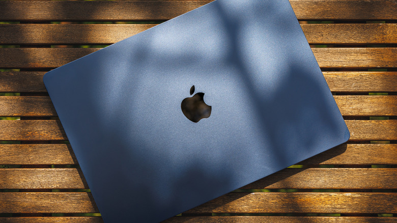 Blue MacBook resting on a wooden outdoor table in direct sunlight, with tree shadows falling across the lid.