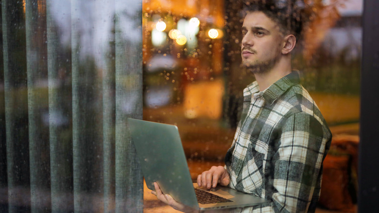Man holding and using a laptop beside a rain-covered window, illustrating wet-weather conditions that can affect outdoor laptop use.