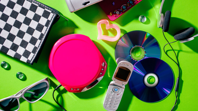 Vintage 2000s technology on a lime green background: a CD player and CDs, wired headphones and a flip phone alongside sunglasses and a plaid bag.
