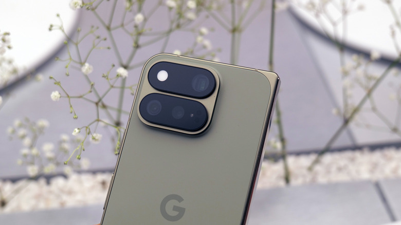 The rear camera layout of the Pixel 11 Pro Fold in Olive, in front of some white flowers.