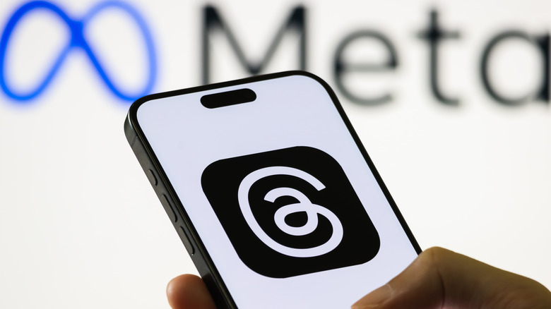 Photo of the Threads logo on a smartphone screen with the Meta logo blurred on a white wall in the background.