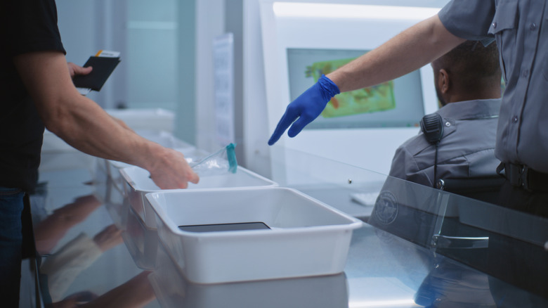 A TSA agent's hand pointing to electronics in a bin at the security checkpoint, a man holding his passport on the opposite side.
