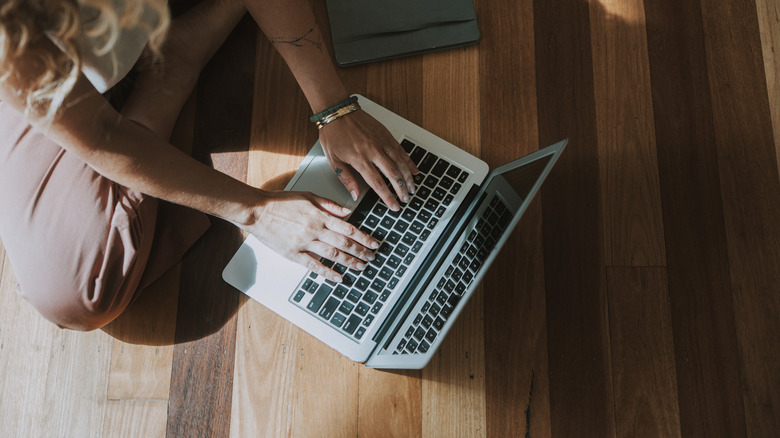 Person using a MacBook on a wooden floor in sunlight.