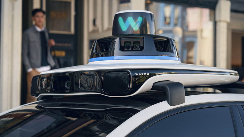 The sensor panel atop a Waymo autonomous car is shown with a passenger in the background