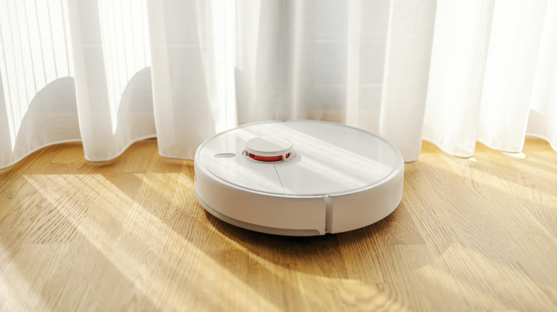 A white robot vacuum cleaner, cleaning near curtains on a wooden floor