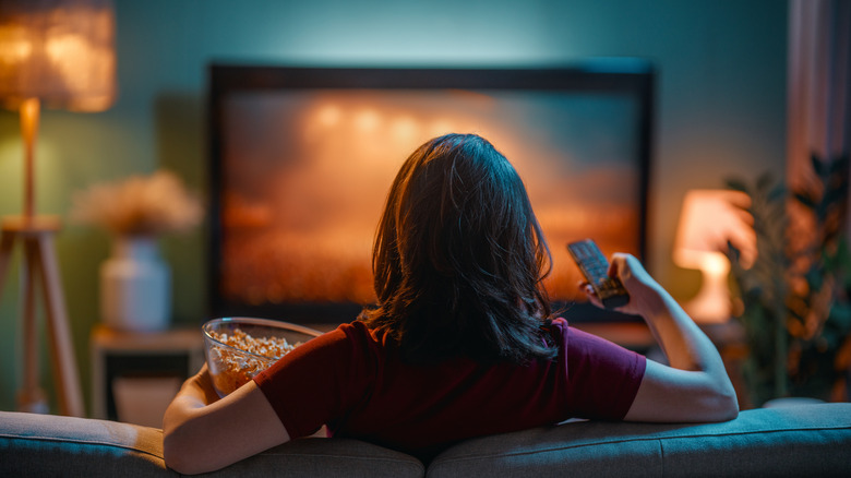 Rear view of a woman sitting on the couch, who raises a TV remote while eating popcorn and watching TV.