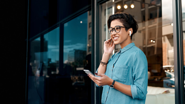 A woman in a blue shirt and glasses holding her phone smiles as she tests her wireless earbuds.
