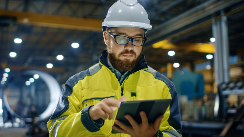 A bearded man in a hard hat and yellow work jacket types on a tablet in the middle of a factory room floor.