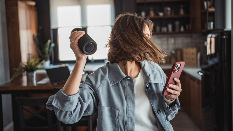 A woman dances in her kitchen, holding a portable speaker and smart phone.
