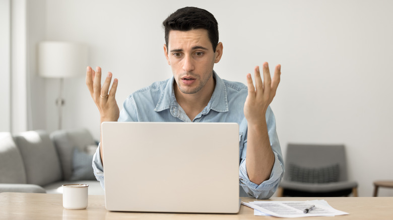 A man looking concerned sitting at a laptop, throwing his hands in the air.