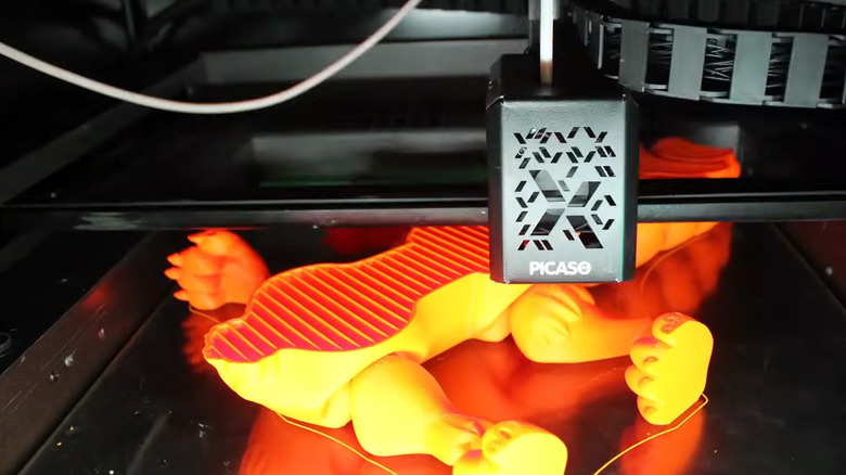 The base of a bright orange creature is layered onto a platform by an FDM 3D printer.