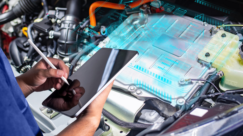 A technician uses a tablet beside an exposed hybrid vehicle powertrain with a digital battery-system graphic overlaid.
