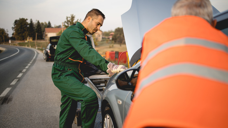 Roadside assistance workers tow a gray car with its hood open on the side of a rural road.