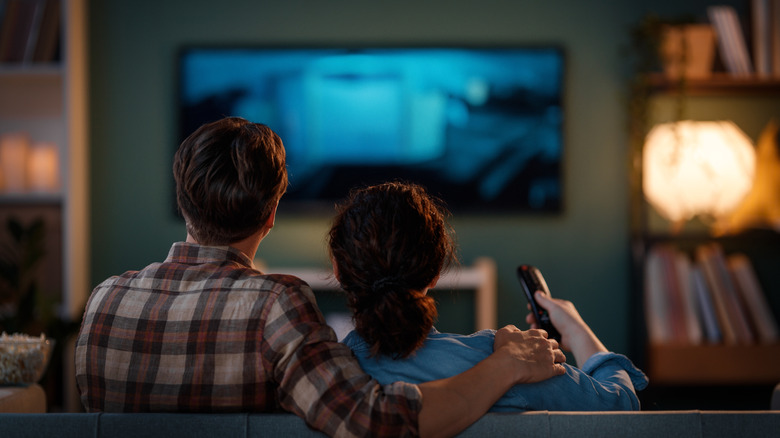 A couple, seen from behind, in blue and red shirts hold each other as they watch television.