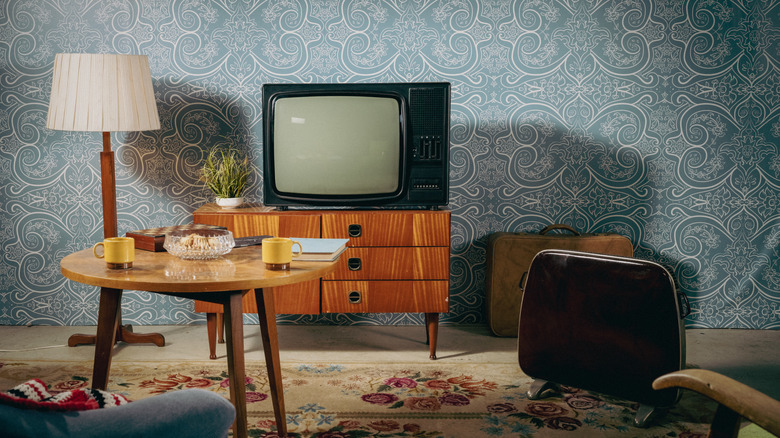 An old timey television set stands atop a chest of drawers in a retro-fitted room with blue wallpaper.
