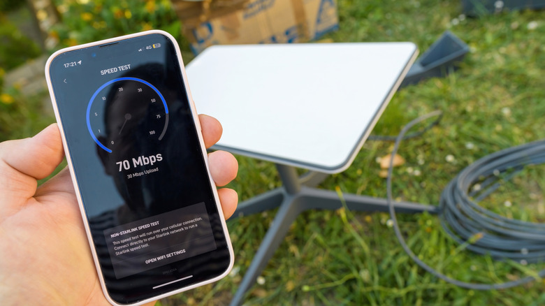 A hand holding a smartphone showing a speed test with a satellite internet dish on the ground.