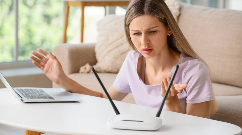 A frustrated woman looking at a wireless router with her laptop on the table.