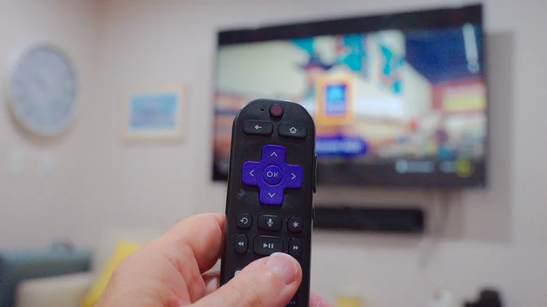Close-up of a hand holding a Roku remote with a TV in the background.