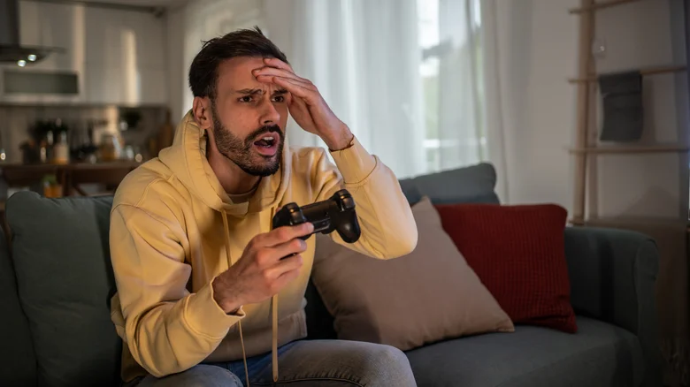 A man sitting on a couch holding a PlayStation controller and looking frustrated.