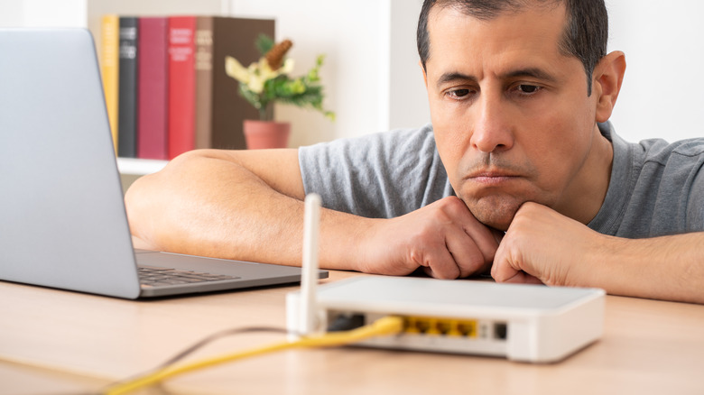 A sullen person, frustrated with their Wi-Fi router while sitting at a desk with a laptop