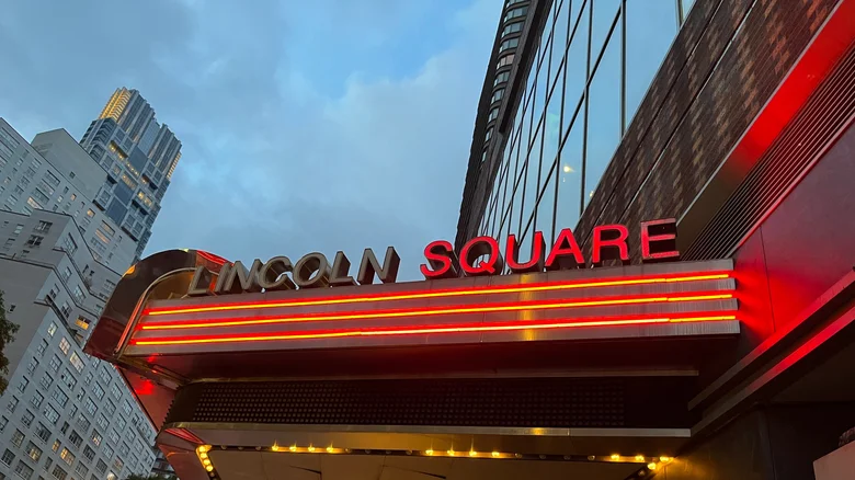 A neon display shows Lincoln Square in bright red letters above a marquee against a cloudy New York City evening.