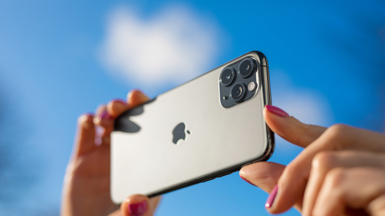 A woman with painted nails snaps a photo with her grey iPhone against a blue sky.