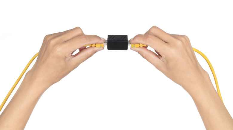 Two hands holding yellow Ethernet cables on either side of a black RJ45 coupler.