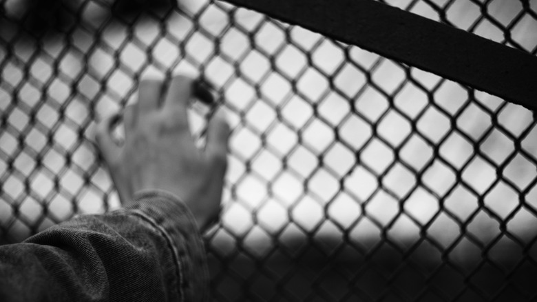 Black and white view of a person's hand against a chain-linked (presumably prison) fence