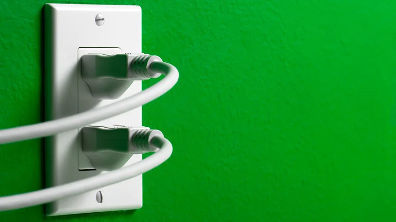 Two white electrical cords plugged into a wall outlet on a green wall.