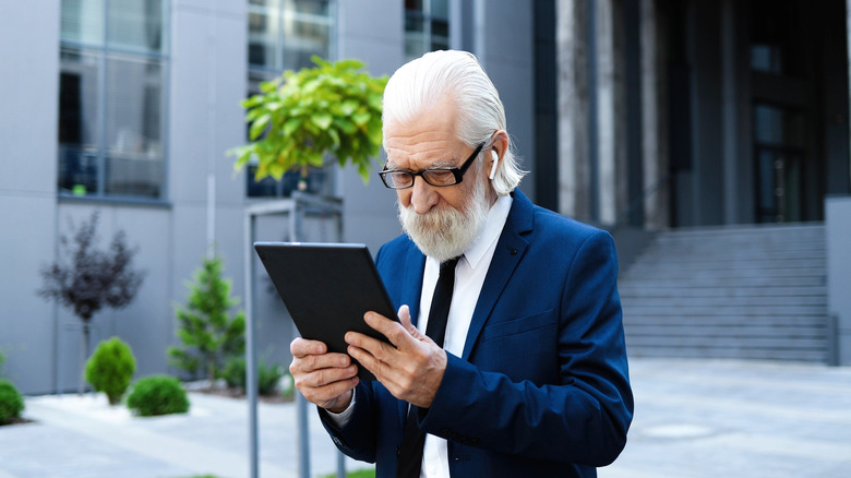 An older,  white bearded man wearing a blue suit listens to his headphones while looking at a tablet.