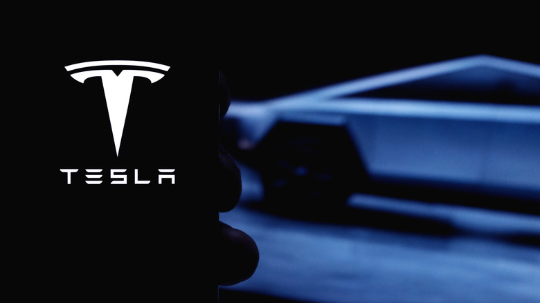 A smartphone displaying the Tesla logo, with a blurred vehicle in the background.