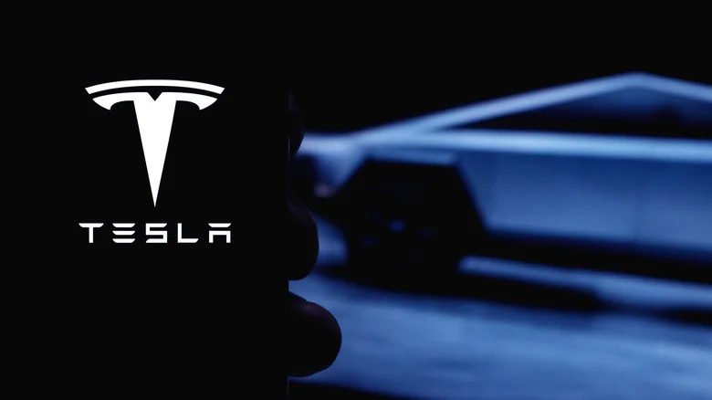 A smartphone displaying the Tesla logo, with a blurred vehicle in the background.