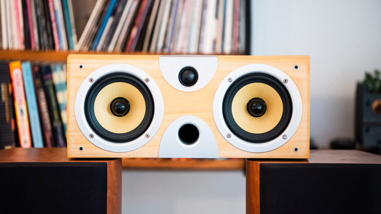 Wooden home speaker with two large yellow drivers and a central tweeter, positioned in front of a shelf of vinyl records and books.