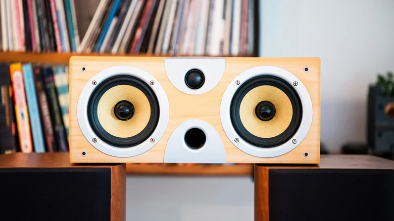 Wooden home speaker with two large yellow drivers and a central tweeter, positioned in front of a shelf of vinyl records and books.