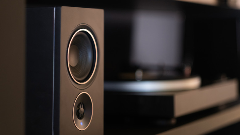 Black bookshelf speaker in a home entertainment setup, with a TV and media console blurred in the background.